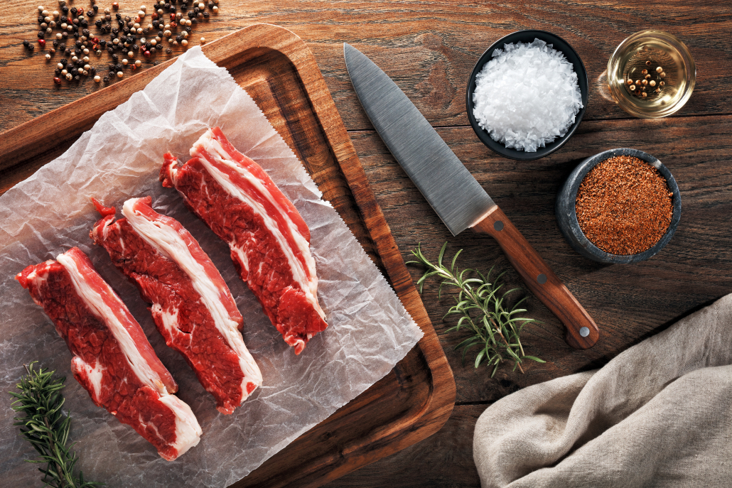 Raw calf short rib on white cooking paper and wooden cutting board. Decorated with herbs, spices and chef's knife. Overhead view.