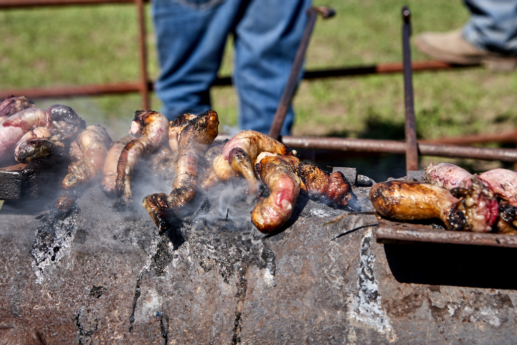 Meat cooking on top of steaming metal grilling device while men lean against metal fence in the background in Flint Hills, Kansas, USA
