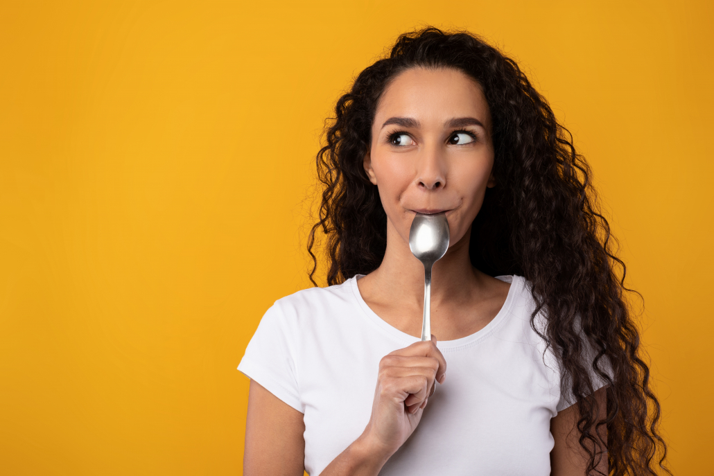 Portrait of excited hungry young woman holding spoon in her mouth dreaming about delicious healthy food, choosing something to eat looking aside at free copy space, yellow orange studio background