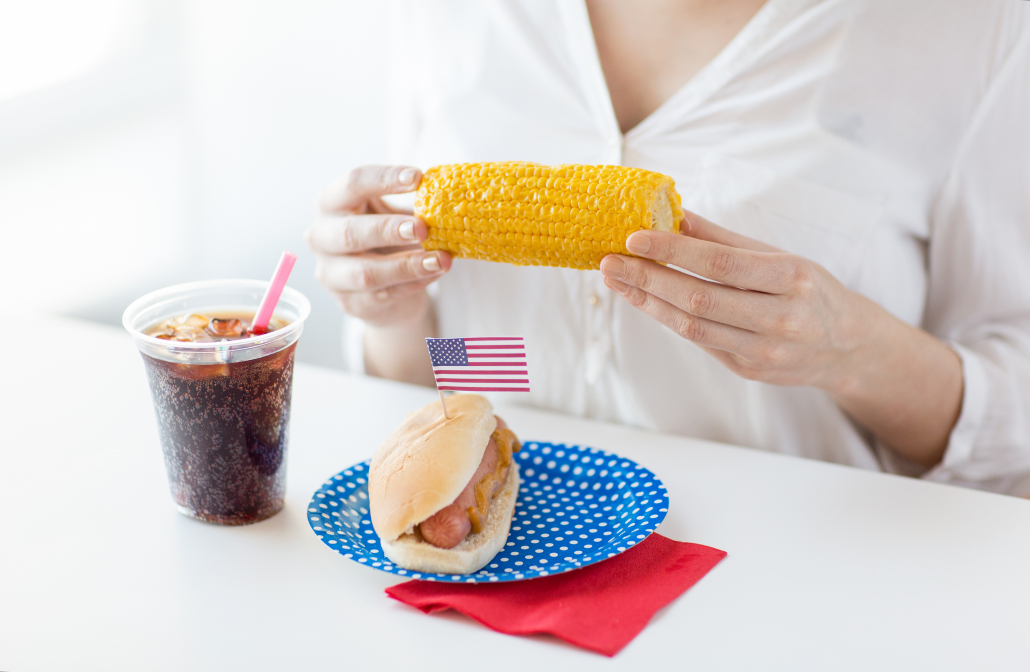 american independence day, celebration, patriotism and holidays concept - close up of woman hands holding corn with hot dog and coca cola drink in plastic cup on 4th july party