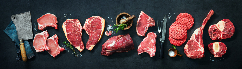 Variety of raw cuts of meat, dry aged beef steaks and hamburger patties for grilling with seasoning and utensils on dark rustic board