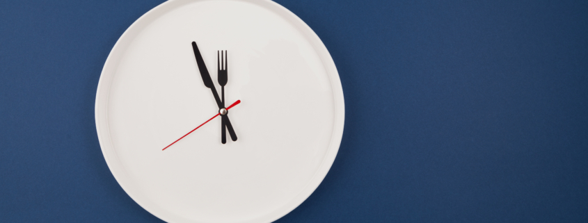 White plate clock on a blue background with copy space. The hands point to 12 o'clock. Interval fasting or autophagy.