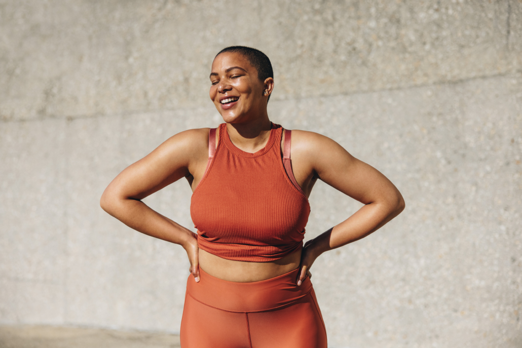 Plus size female model in sportswear smiling with her hands on hips. Positive woman relaxing after workout outdoors.