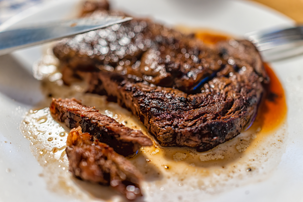 Closeup of knife slicing cutting cooked medium rare well done grilled grass-fed ribeye rib eye meat beef steak on white plate