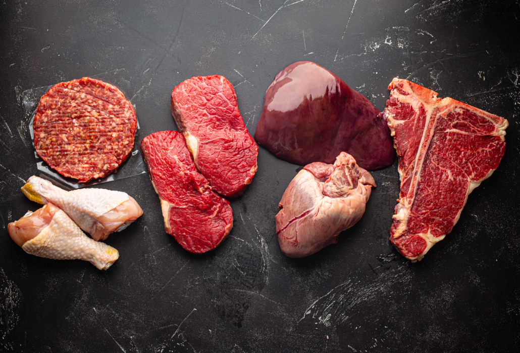 Selection of assorted raw meat food for zero carb carnivore diet: uncooked beef steak, ground meat patty, heart, liver and chicken legs on black stone background from above